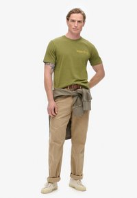 Olivengrøn bomulds t-shirt med "superdry" logo i guld, parret med beige kordfløjspants. Beige sneakers og en grå sweatshirt bundet om livet.