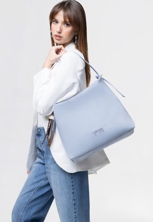 Joven mujer con blazer blanco y vaqueros llevando un gran bolso de hombro azul claro con el logo "V°78" sobre fondo liso.