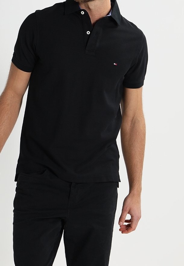 Homme portant un polo noir à manches courtes avec des boutons et un petit logo, associé à un pantalon noir, debout devant un fond uni.