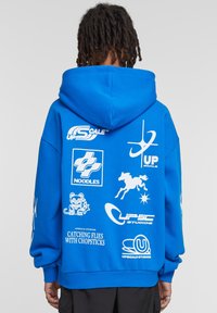 Sweatshirt cu glugă albastră, având logo-uri grafice mari și albe, inclusiv text și simboluri, pe spate. Materialul pare moale, cu o croială lejeră.