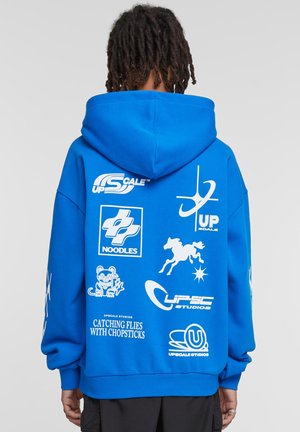 Blue hooded sweatshirt featuring large white graphic logos, including text and symbols, on the back. Fabric appears soft with a relaxed fit.