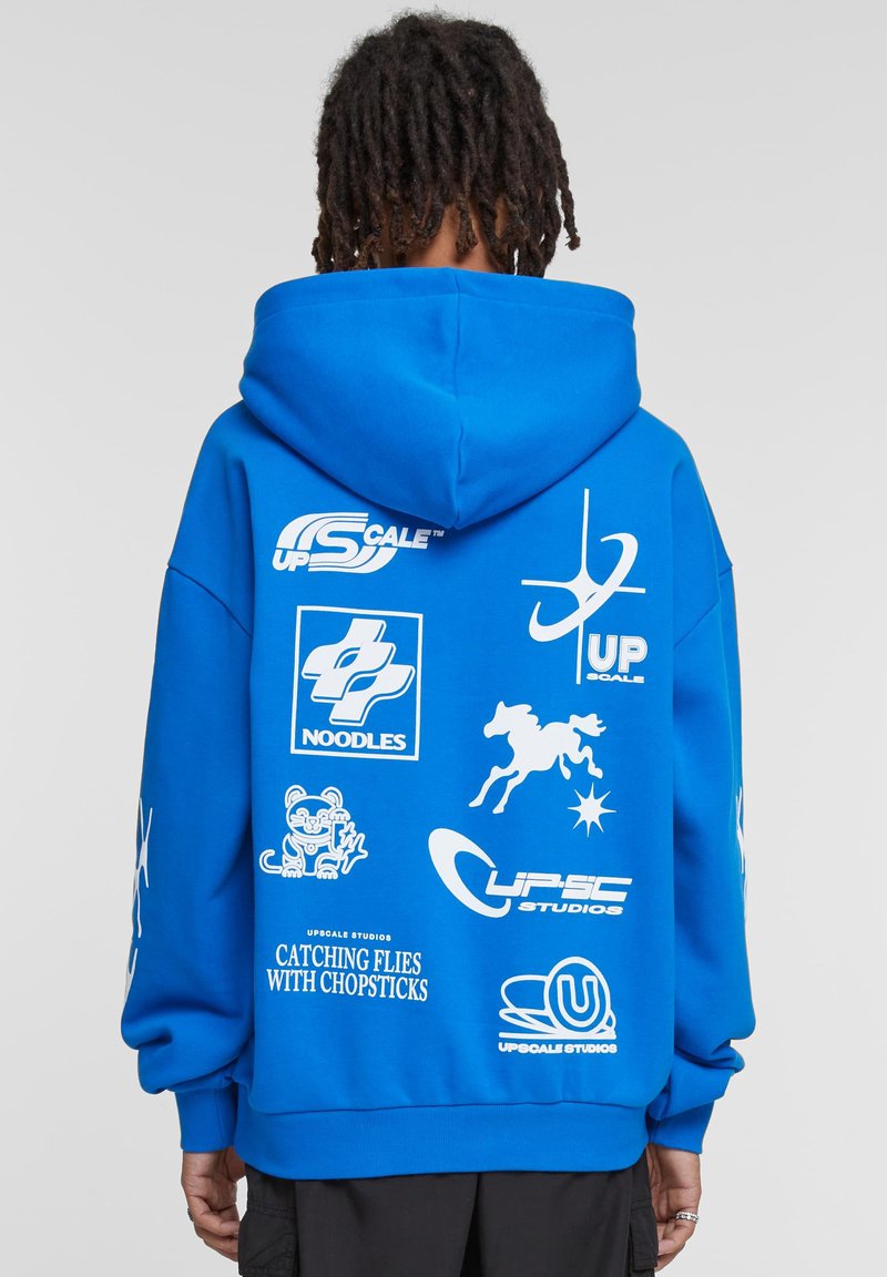 Sweatshirt cu glugă albastră, având logo-uri grafice mari și albe, inclusiv text și simboluri, pe spate. Materialul pare moale, cu o croială lejeră.