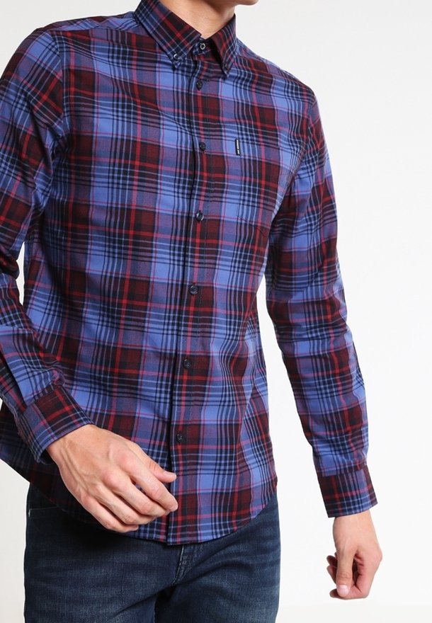 Homme portant une chemise à carreaux rouge et bleu à manches longues boutonnée avec un jean foncé, visible du cou aux hanches sur un fond blanc.