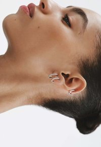 Pendientes de oreja de plata con un diseño curvado, que presentan acentos detallados. Textura suave, posicionados en la oreja en un ángulo ascendente.