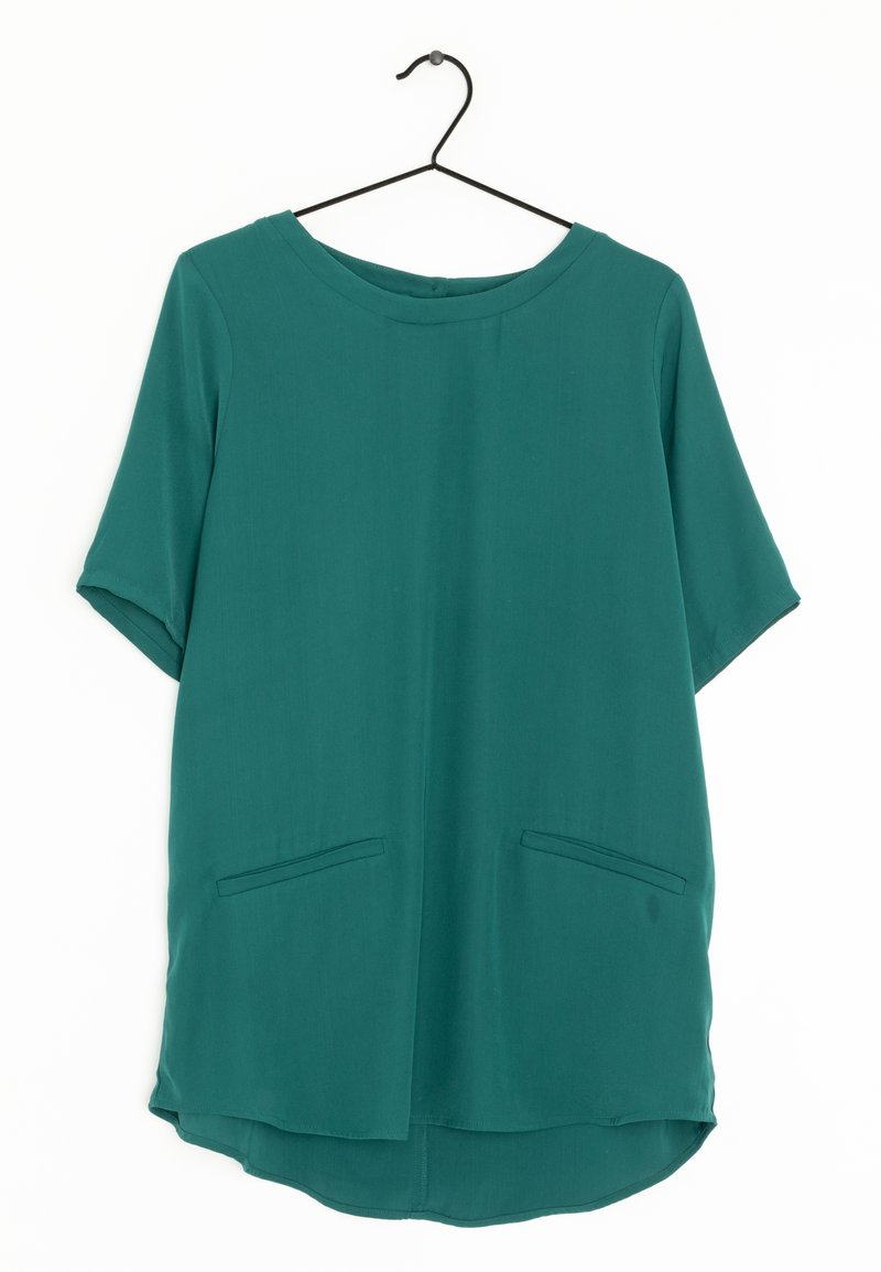 Blouse turquoise à manches courtes avec un col rond, deux poches avant et un ourlet arrondi. Tissu lisse avec une texture légère.