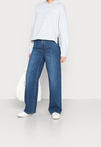 Persona che indossa un hoodie corto di colore blu chiaro, jeans a gamba larga blu, sneaker bianche, mentre tiene una grande borsa bianca contro uno sfondo semplice.