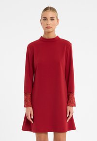 Rotes Kleid mit langen Ärmeln und Spitzenakzenten an den Manschetten. Hergestellt aus einem glatten Stoff, hat es einen hohen Ausschnitt und eine A-Linie Silhouette.