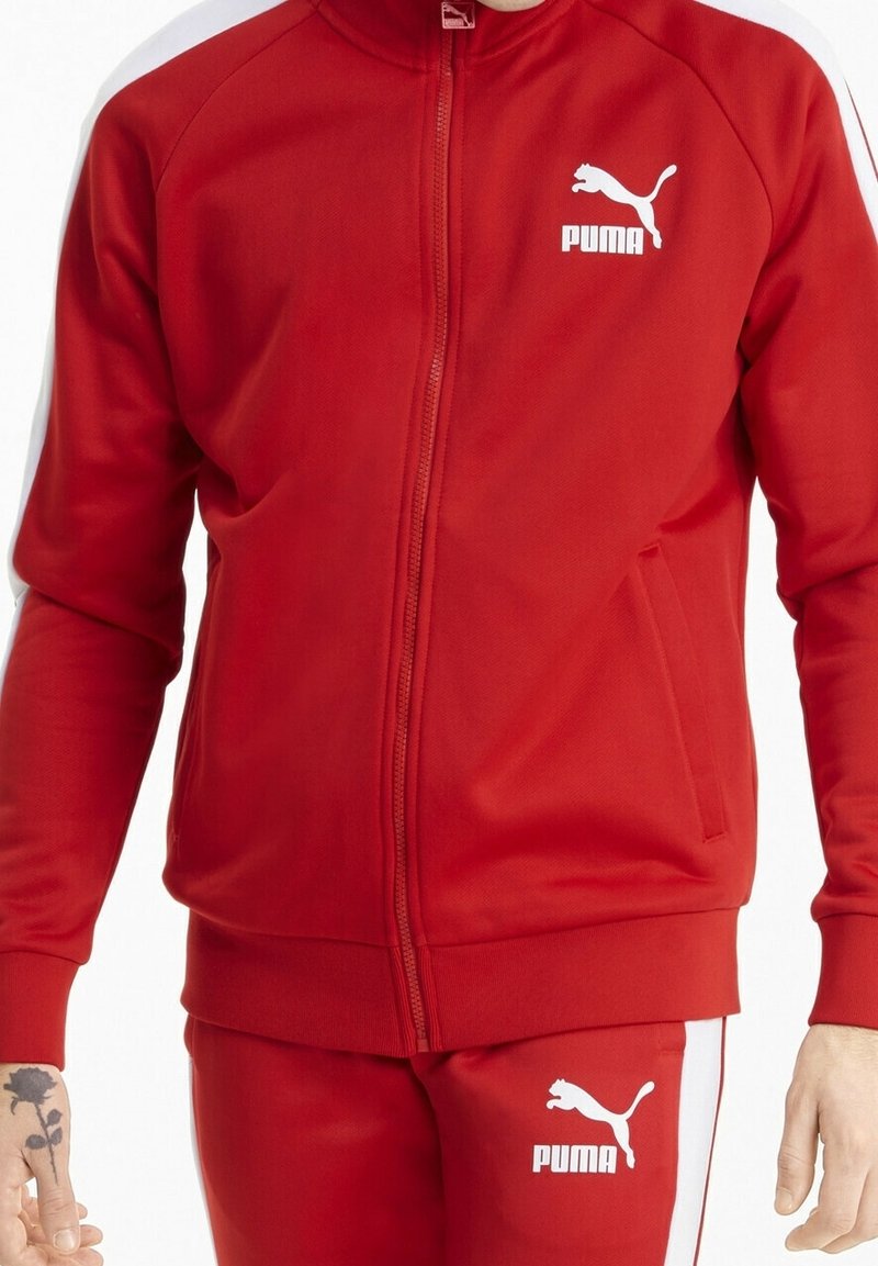 Giacca rossa con zip realizzata in tessuto liscio. Presenta il logo bianco Puma sul petto, polsini elasticizzati e tasche laterali. Nessun motivo visibile.