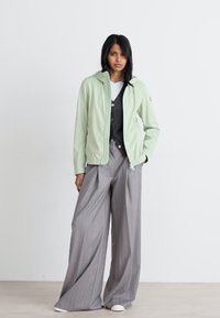 Világoszöld széldzseki, elől cipzárral és kapucnival, egy szürke kardigánra és széles, csíkos szürke nadrágra rétegezve, fehér cipőkkel kiegészítve.