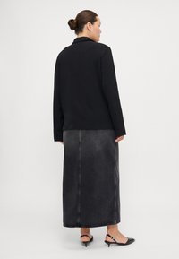 Blazer noir à manches longues sur une jupe en denim noire ajustée et mi-longue ; texture lisse, délavage gris subtil, détail de couture au dos, et des talons.
