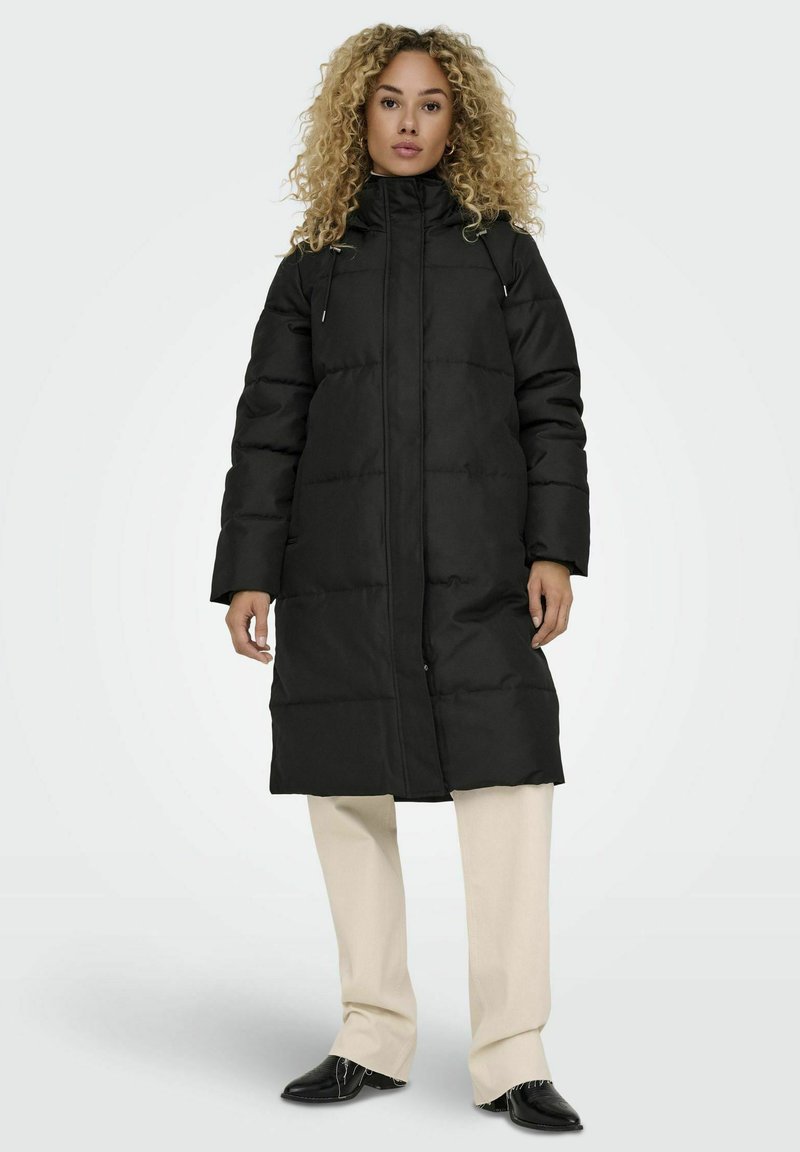 Manteau noir rembourré avec un col haut, doté d'une fermeture éclair à l'avant et de deux poches latérales. Porté sur un pantalon crème et des chaussures noires.