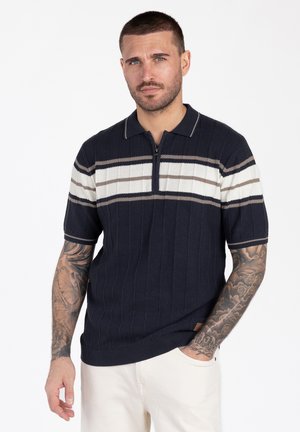 Mężczyzna o krótkich włosach i tatuażach, ubrany w granatową dzianinową koszulkę polo z białymi i beżowymi paskami, stojący z jedną ręką w kieszeni na białym tle.