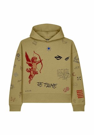 Jonge vrouw draagt een beige hoodie met prints van cupido, ogen en tekst, gecombineerd met zwarte cargobroek en witte sneakers, staand tegen een witte achtergrond.
