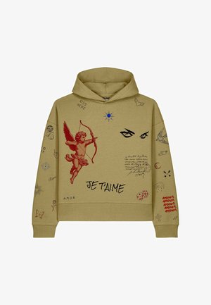 Jeune femme portant un sweat à capuche beige avec des motifs cupidon, yeux et texte, associée à un pantalon cargo noir et des baskets blanches, debout sur fond blanc.