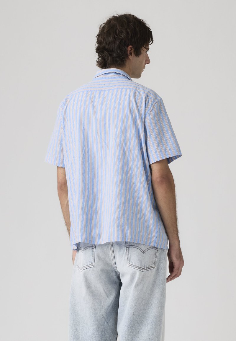 Hombre con cabello rizado oscuro que lleva una camisa de manga corta a rayas azul claro y beige y jeans de mezclilla claros, visto de espaldas.