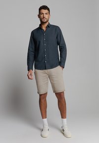 Donkerblauwe overhemden van stof, gecombineerd met beige shorts. De outfit heeft opgerolde zoom en witte sneakers met subtiele accenten.