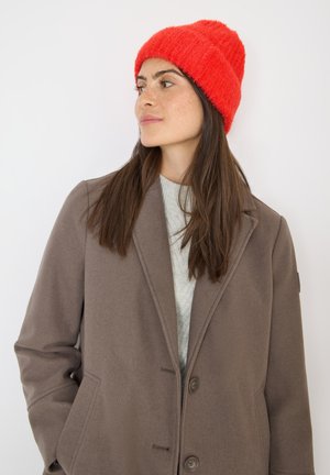 Brun uldfrakke med klapkrave, to knapper og sidelommer. Rød fuzzy strikket hue oven på langt, mørkt hår. Lys grå sweater under.