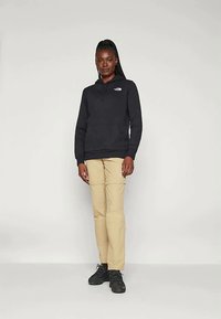 Γυναίκα ντυμένη με μαύρη κουκούλα, μπεζ παντελόνι και μαύρα αθλητικά παπούτσια, στέκεται μπροστά από ένα απλό φωτεινό φόντο με χαλαρή στάση.