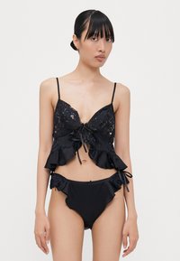 Melnas divdaļīgas peldkostīms ar mežģīņu topu, kas rot�āts ar volāniem, un atbilstošām bikini biksītēm ar volānu apdari un sānu saitēm.