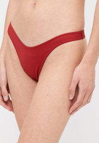 Pantaloni de baie roșii, cu o talie subțire, cu un model cu talie joasă și o textură netedă. Oferă o acoperire minimă și detalii de cusătură curate.