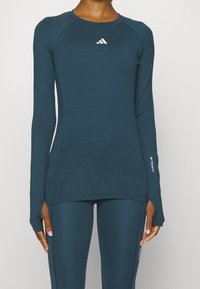 Person som bär en mörk teal Adidas långärmad träningsskjorta och matchande leggings med tumhål och synliga logotyper på bröstet och ärmarna.
