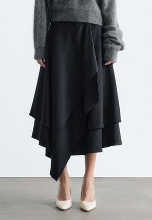 Femme portant une jupe noire asymétrique à couches, un pull gris pelucheux et des escarpins blancs à bout pointu, debout sur un sol gris.