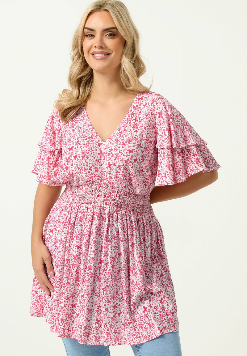 Lächelnde Frau mit blondem Haar, die ein rosa-weißes, geblümtes V-Ausschnitt-Kleid mit flatternden Ärmeln und geraffter Taille trägt, steht mit einer Hand in der Hüfte.