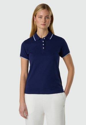 Mujer con cabello largo y rubio, vestida con una camisa polo azul marino de manga corta con ribete blanco y pantalones blancos, de pie frente a un fondo neutro.