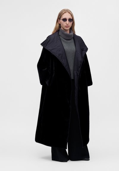 Γυναίκα που φοράει μαύρο oversized παλτό, γκρι πουλόβερ με ζιβάγκο, φαρδιά μαύρη παντελόνια και μικρά στρογγυλά γυαλιά ηλίου, στέκεται μπροστά σε απλό φόντο.