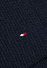 Pull en tricot côtelé bleu marine avec un petit patch logo rouge, blanc et bleu. La texture semble douce et tissée, avec des lignes verticales prononcées.