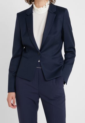 Femme portant un blazer bleu marine cintré à un seul bouton, par-dessus un chemisier blanc à col volanté et un pantalon bleu marine assorti.