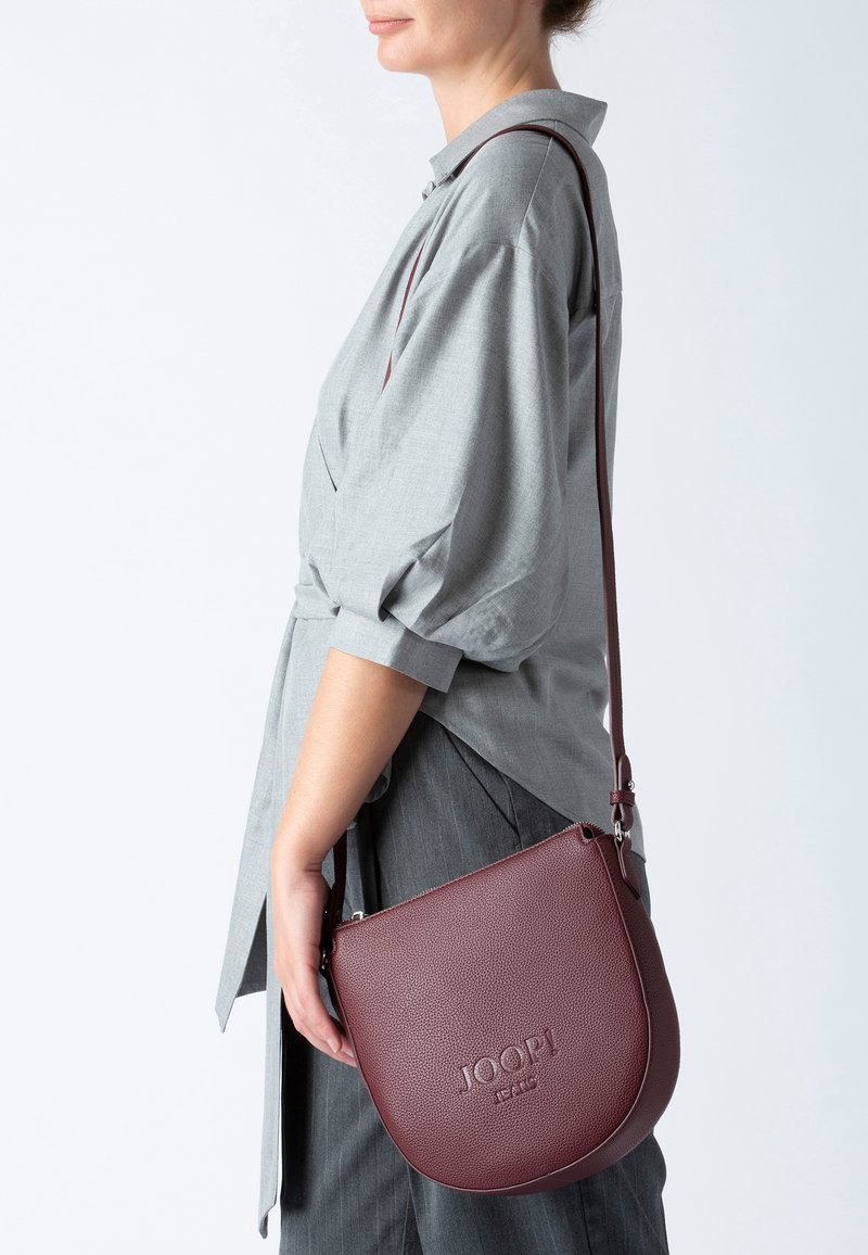 Vinröd läder crossbody-väska med texturerad yta, dragkedjestängning och justerbar rem. Har prägling av logotyper på framsidan.