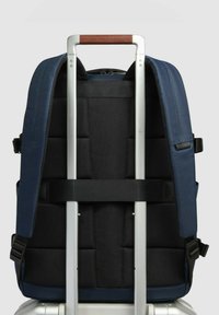 Zaino blu navy con schienale e spallacci imbottiti, dotato di manico da trolley in alluminio argento e impugnatura in pelle marrone.