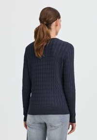 Maglione lavorato a maglia blu navy con motivo testurizzato, maniche lunghe, polsini e orlo a coste. Indossato sopra jeans grigio chiaro, visto da dietro.