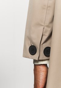 Manga de un abrigo beige con dos grandes botones negros sobre una manga blanca arremangada y una porción de la muñeca y la mano de una persona.