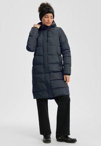 Moteris, vilkinti ilgą juodą pūkinę striukę, juodas kelnes, juodus aulinukus ir juodą galvos juostą, stovi prieš paprastą šviesų foną.