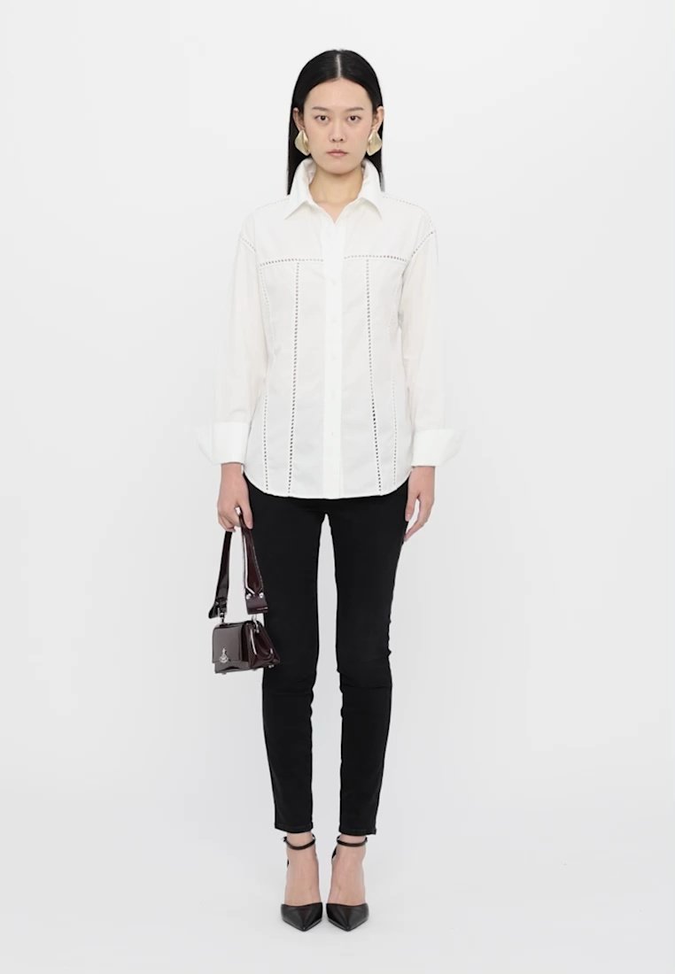 Chemise blanche à manches longues avec des coutures décoratives, jeans skinny noirs, escarpins noirs à bout pointu et un petit sac à main sombre.