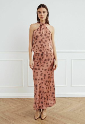 Nő ujjatlan, nyakpántos felsőt és a hozzá illő, barna absztrakt foltokkal díszített bokáig érő szoknyát visel, fából készült padlón áll, fehér fal előtt.