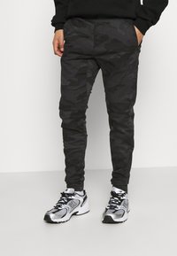 Černé maskáčové joggery s úzkým střihem, hladkou texturou a elastickým pasem, doplněné o šedé a černé sportovní boty.