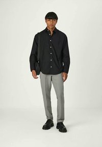 Homem a usar uma camisa preta de botão, calças cinza claro, sapatos pretos e a carregar uma mala de ombro preta, num fundo neutro.
