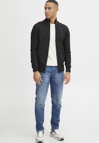 Svart texturerad zip-up tröja med stående krage, bärs över en vit T-shirt, tillsammans med ljusblå jeans och grå sneakers.