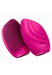 Brosse de nettoyage en silicone rose avec des poils texturés d'un côté et un design ondulé de l'autre, comportant deux boutons et le logo de la marque.