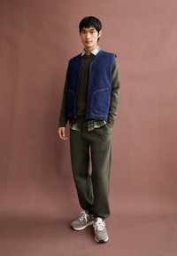Denim vest over een olijfgroen trui, gelaagd met een geruit shirt, gecombineerd met olijfgroene sweatpants en grijze sneakers. Effen bruine achtergrond.