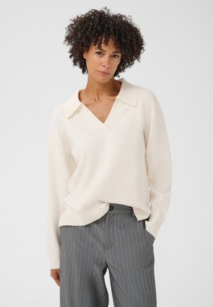 Femme aux cheveux bouclés portant un haut à manches longues écru et un pantalon gris à fines rayures, debout devant un fond clair et uni.