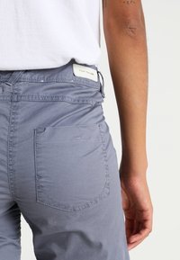 Lichtgrijze katoenen broek met een gestikte achterzak en een label met het merk "TOM TAILOR" op de tailleband. Gladde textuur, casual ontwerp.