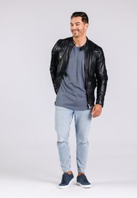 Svart läderjacka med kviltade detaljer, grå t-shirt, slitna ljusblå jeans och marinblå sneakers. Stående mot en neutral bakgrund.
