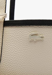 Borsa in pelle di colore crema con bordo nero, dotata di un piccolo logo di coccodrillo embossato di colore chiaro sulla superficie.
