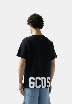 Persona con capelli ricci vista da dietro, che indossa una t-shirt nera con grandi lettere bianche "GCDS" nella parte bassa della schiena e pantaloncini di jeans azzurri chiari.