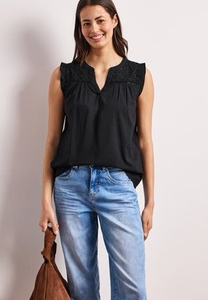 Lachende vrouw die een mouwloze zwarte top met kanten details draagt en lichtblauwe spijkerbroek, houdend een bruine suède handtas aan haar zijde.