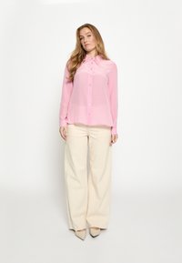 Lyserød, langærmet bluse med knapper foran, kombineret med brede elfenbensfarvede bukser og spidse beige hæle. Glat, gennemsigtigt stof.